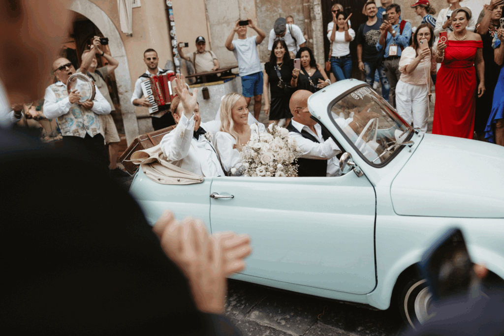 Just married couple waving from a vintage mint Fiat 500 during their Italian destination wedding celebration, surrounded by cheering guests
