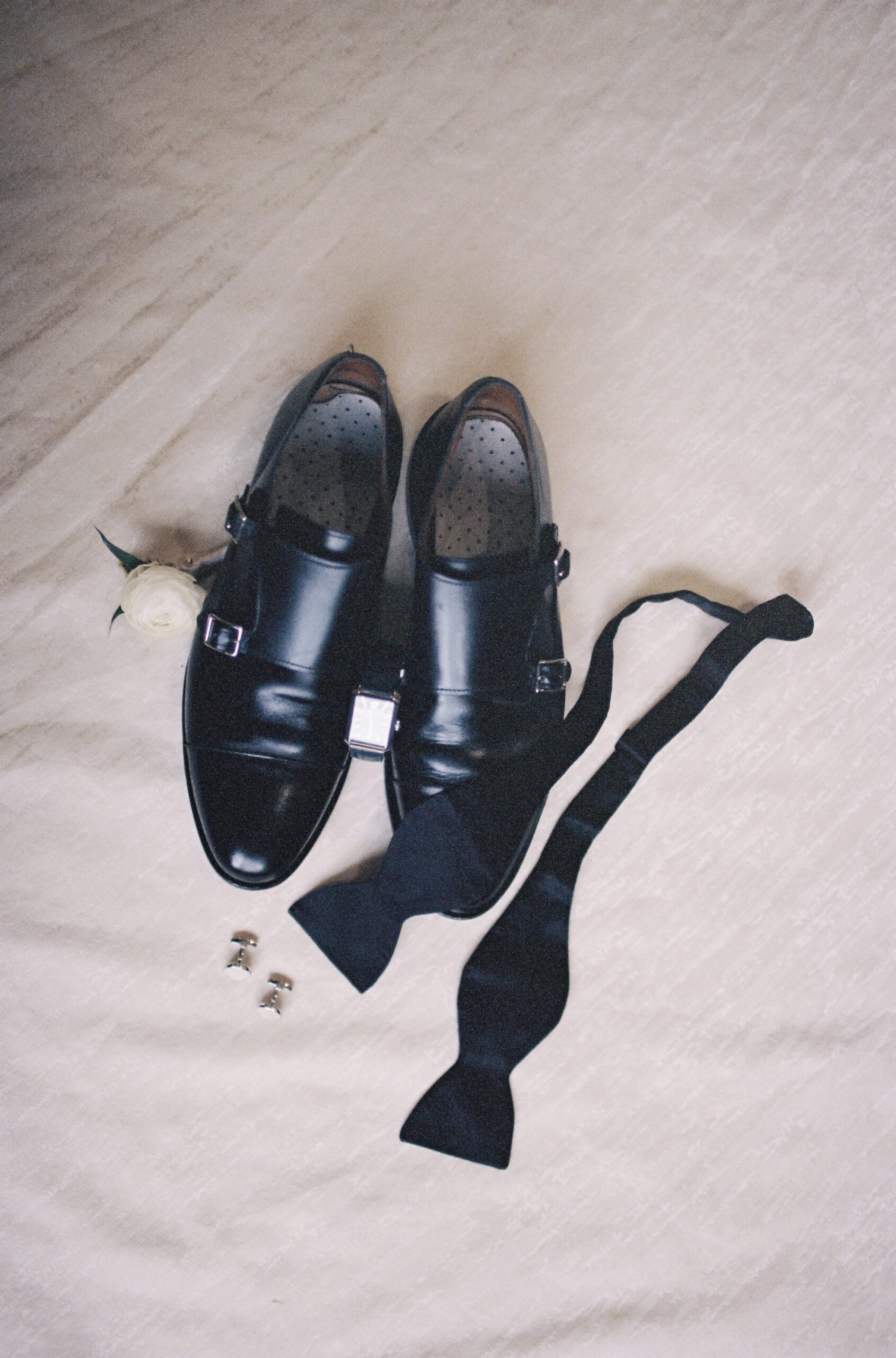 Flat lay of black groom’s dress shoes, untied bow tie, cufflinks, and boutonnière on wedding day, captured on 35mm film