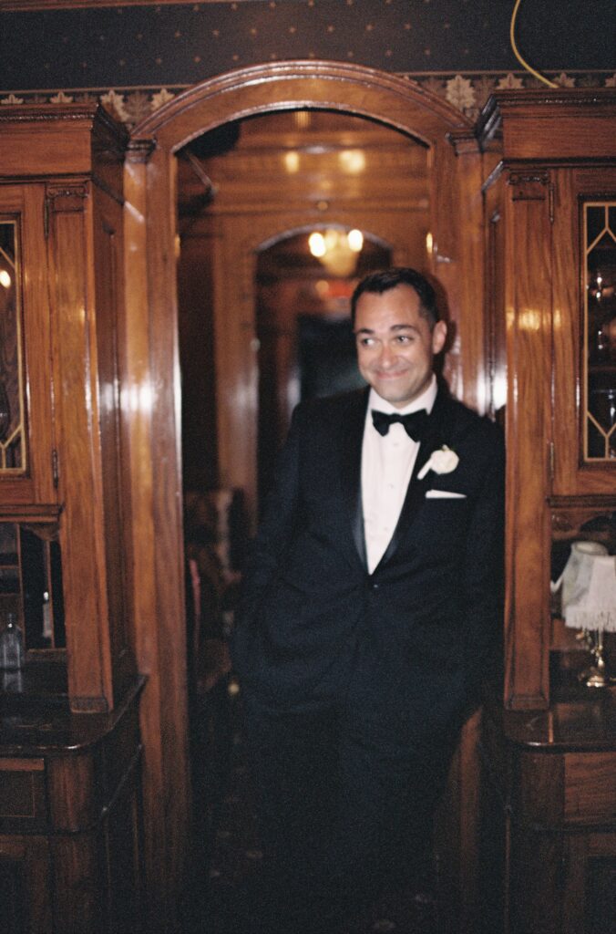 Groom in a tuxedo leaning against a doorway inside a vintage train car.