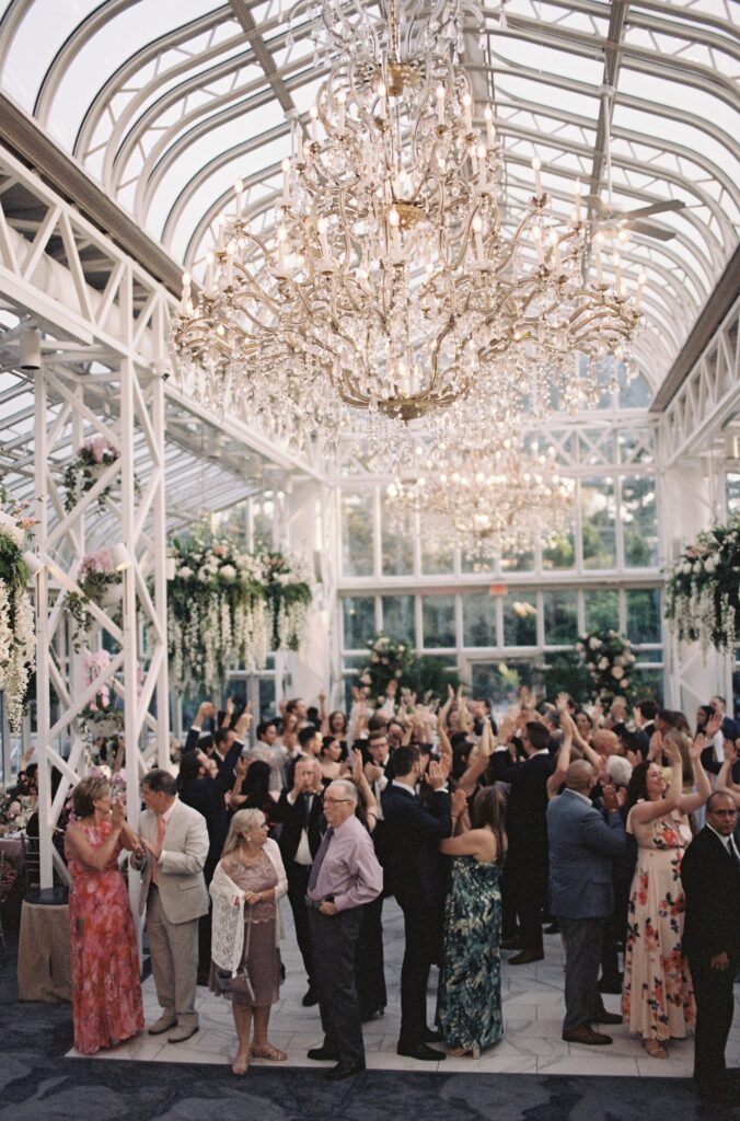 A crowd of wedding guests raising their hands in celebration beneath crystal chandeliers.