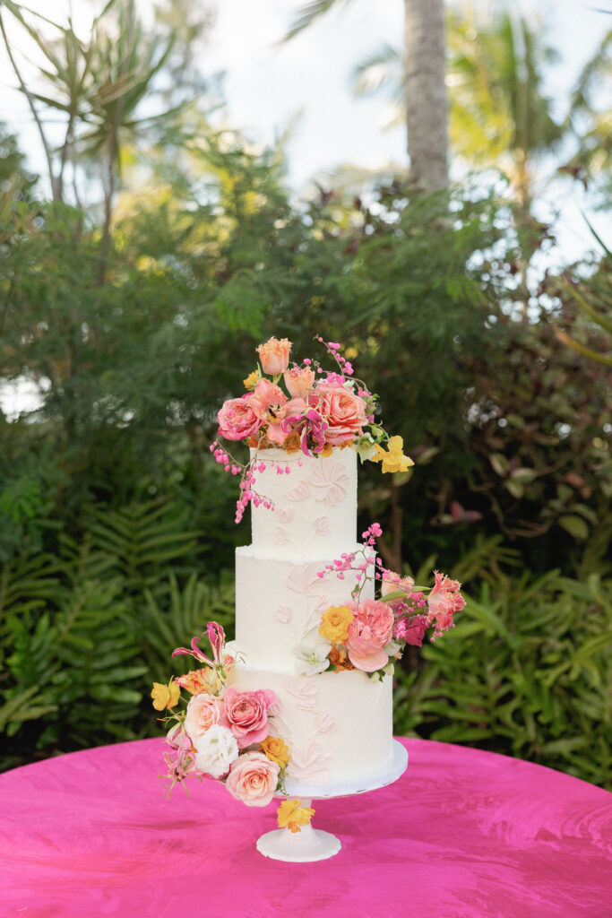 Three-tier white wedding cake with bright tropical flowers on a pink table at Loulu Palm in Hawaii.