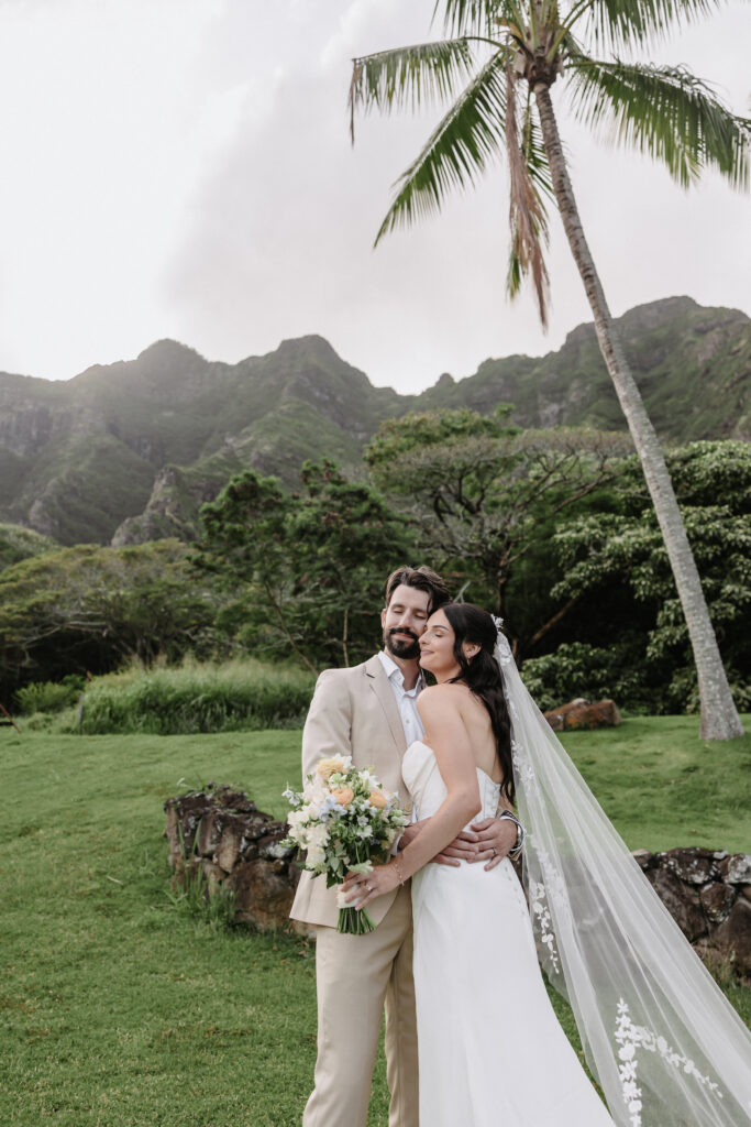 Bride and groom embrace with lush mountains behind them at Kualoa Ranch Paliku Gardens in Oahu, Hawaii