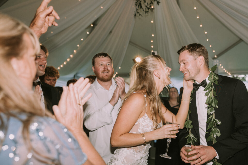 Bride and groom laughing and holding drinks during their wedding reception at Kualoa Ranch Paliku Gardens in Hawaii, surrounded by cheering guests under a draped tent with string lights.