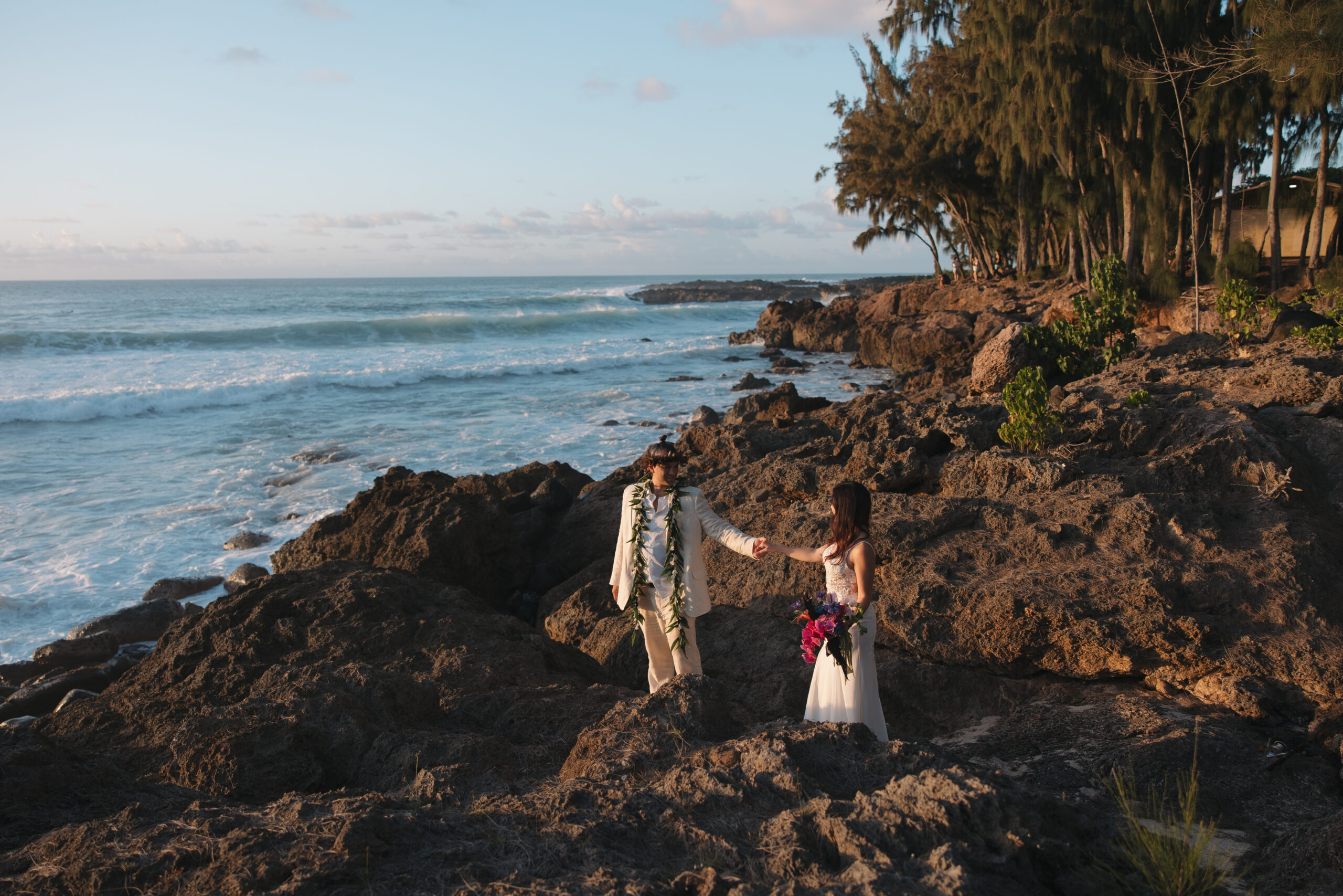 Bride and groom holding hands during golden hour at Sunset Ranch, a scenic coastal wedding venue in Oahu, Hawaii, surrounded by rocky cliffs and ocean waves.