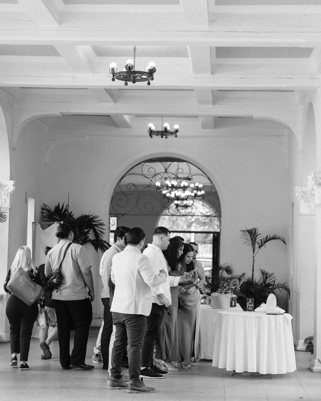 Wedding guests gathered around a welcome table inside a historic villa with arched entryway and chandeliers, photographed in black and white