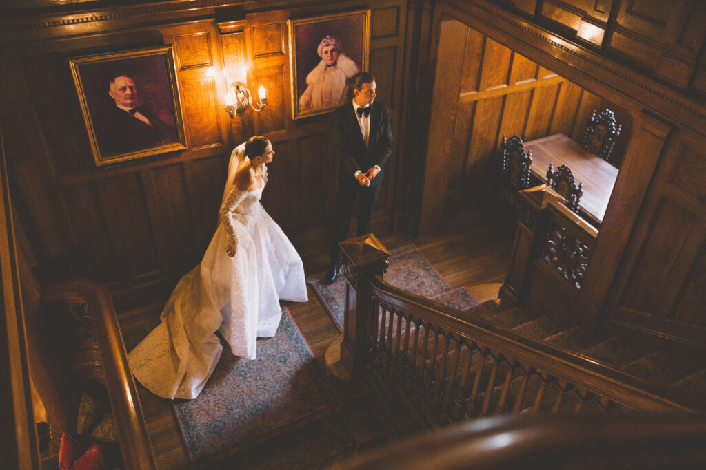 Bride and groom standing on a grand wooden staircase inside a historic European villa, captured in warm candlelit tones