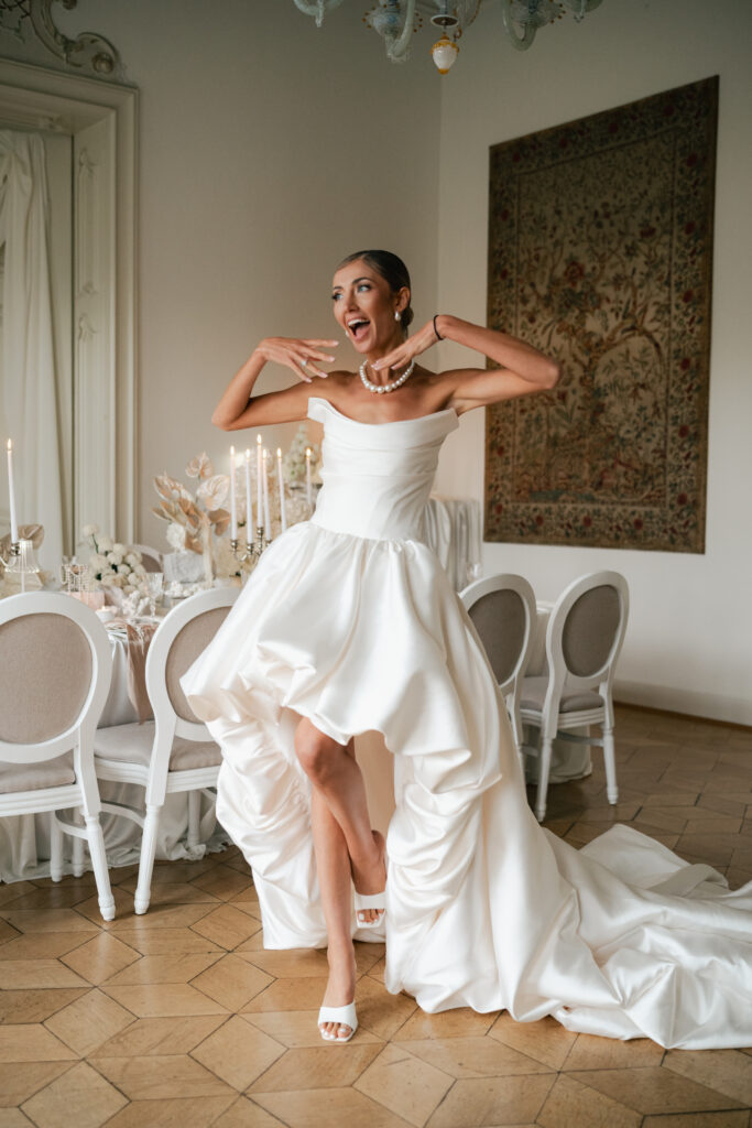 Bride in a strapless white wedding gown laughing and adjusting her necklace inside an elegant European reception room with candlelit tables and classic white chairs.