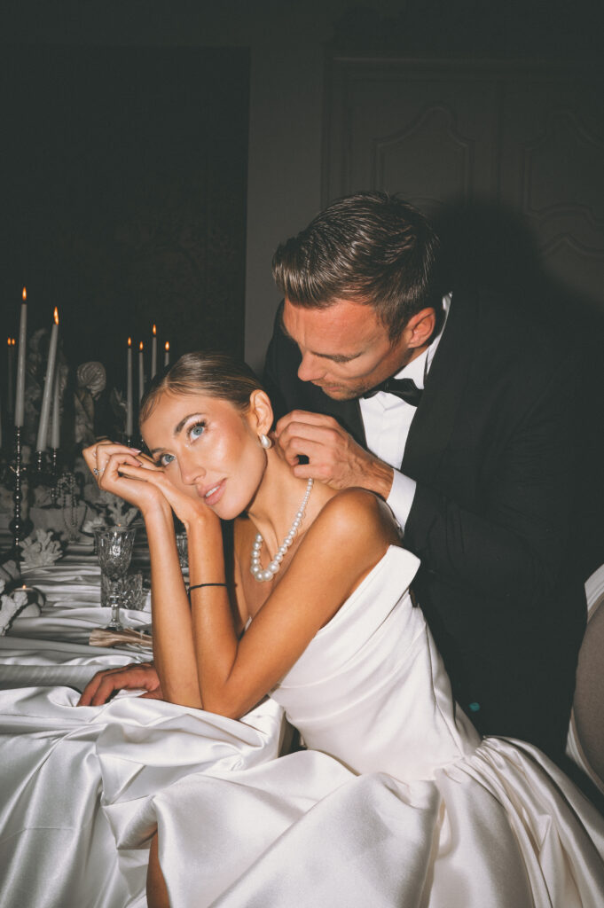 Bride seated at a candlelit reception table while the groom leans in affectionately behind her, showcasing an intimate luxury wedding moment with pearls and soft evening light.