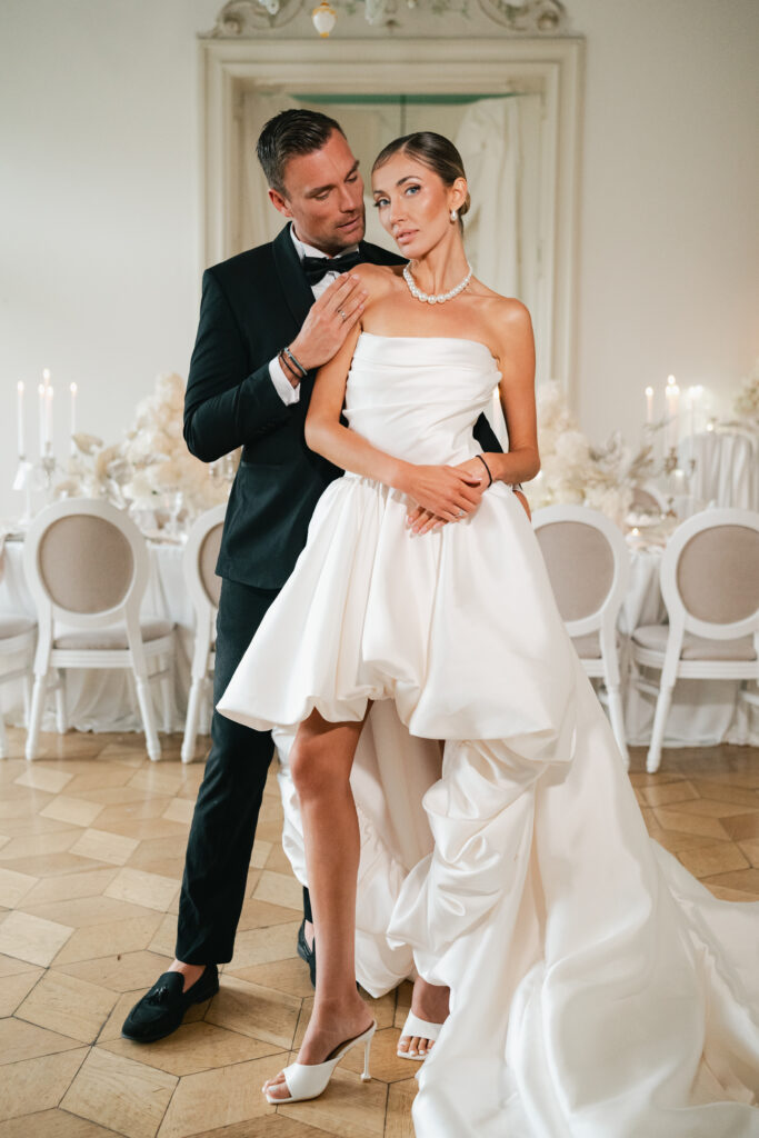 Bride and groom standing together in a refined wedding reception space, the groom embracing the bride from behind as her modern high low wedding dress flows across the tiled floor.
